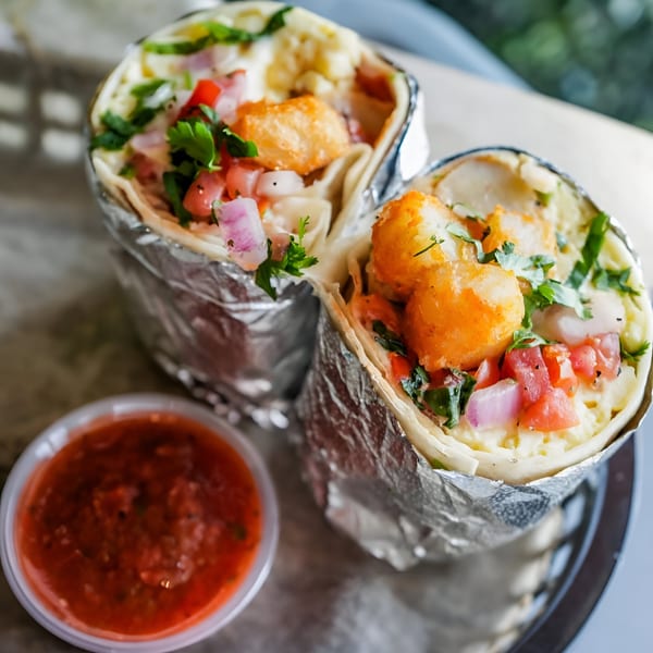 Two foil-wrapped Crush Taco burritos filled with tater tots, pico de gallo, and sauce, with a side of salsa.
