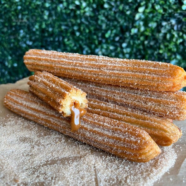 Sugar-dusted churros, one broken open to reveal dulce de leche filling, on brown paper with green background.